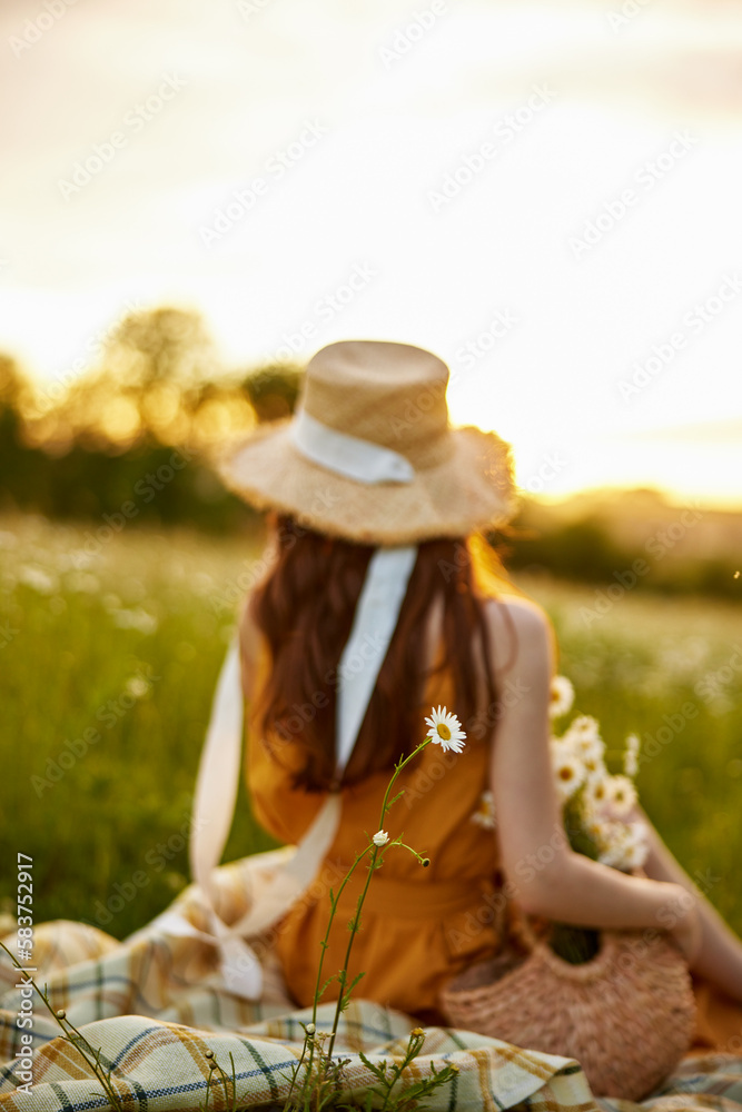 a woman in a wicker hat sits with her back to the camera in a chamomile field on a checkered plaid with a basket of flowers in her hands and looks at the sunset