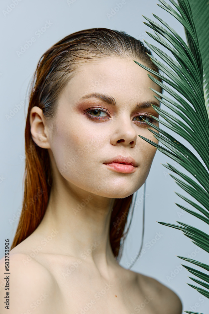a close beauty portrait of a beautiful woman standing on a light background holding a palm leaf near her face, looking into the camera. Vertical photo without retouching of problem skin a close beauty portrait of a beautiful woman standing on a light background holding a palm leaf near her face, looking into the camera. Vertical photo without retouching of problem skin