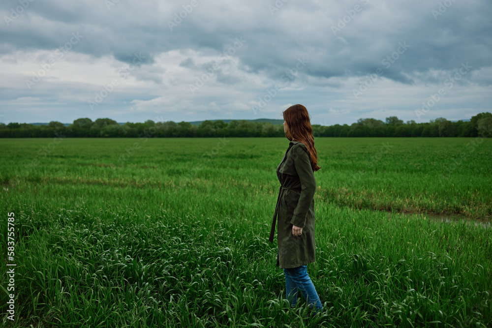 a woman with beautiful, long, red hair stands with her back to the camera in a green field in rainy, spring weather in a long raincoat