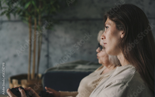 Side view of a woman with her mother playing games on TV, sitting on cozy sofa. Healthy activity. Happy cheerful family. Happy lifestyle. Family home leisure.