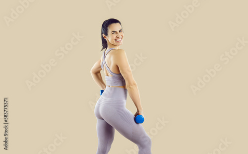 Fototapeta Naklejka Na Ścianę i Meble -  Attractive positive Caucasian woman fitness trainer smiling looking at camera doing sports exercises with mini dumbbells to maintain beautiful body and healthy lifestyle stands on beige background