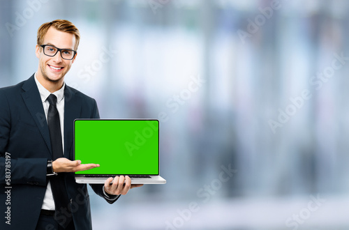 Business man in glasses spectacles, black suit show laptop with empty mockup green chroma key screen, on blurred office background. It expert, technician repair service, tech support maintenance