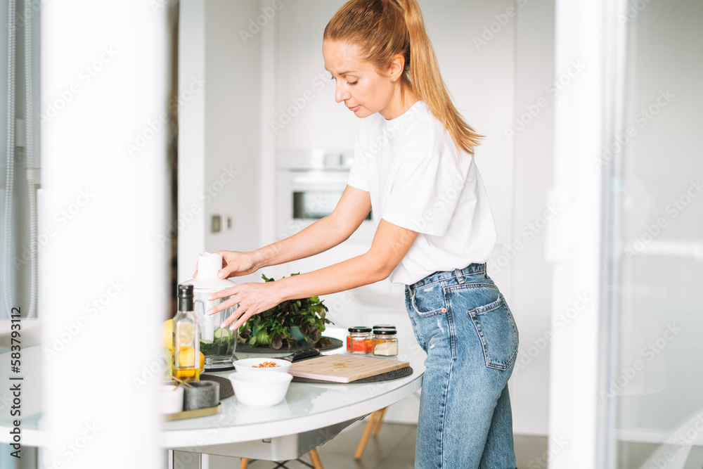 Young slim woman in white t-shirt and blue jeans cooking smoothie healthy food in kitchen at home Young slim woman in white t-shirt and blue jeans cooking smoothie healthy food in kitchen at home