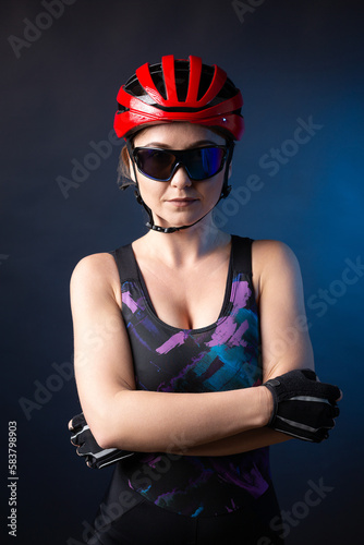 A young female cyclist wearing a safety helmet and glasses, dressed in a bib shorts poses against a black background in the studio. © Budjak Studio