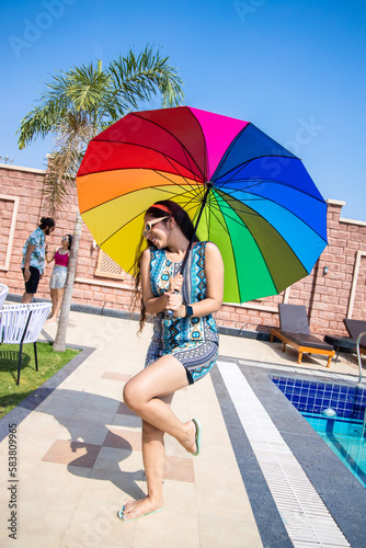 Beautiful young indian woman wearing sunglasses holding rainbow color umbrella in hot summer day. Vacation and holidays.