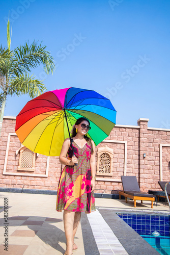 Beautiful happy young indian woman wearing sunglasses holding rainbow color umbrella in hot summer day. Vacation and holidays.