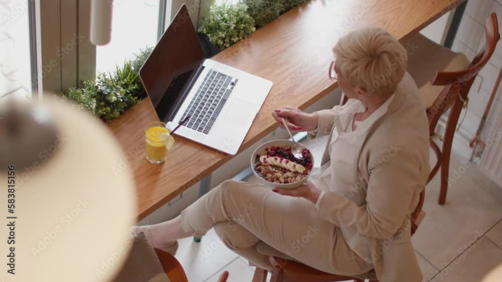 Senior woman using mobile phone while working on the laptop and having healthy breakfast in the cafe