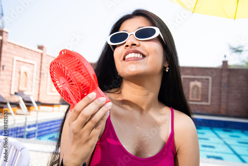 Hot Summer, Young indian girl wear sunglasses enjoying air flow portable electric fan cooling her body outdoor sunny day.  photo