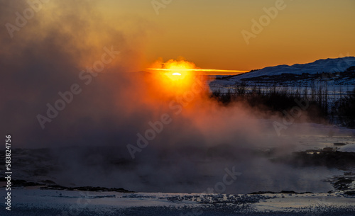 Iceland's great Strokkur geyser just before erupting with mist and smoke backlit with the orange sunset sun behind it. Volcanic activity.
