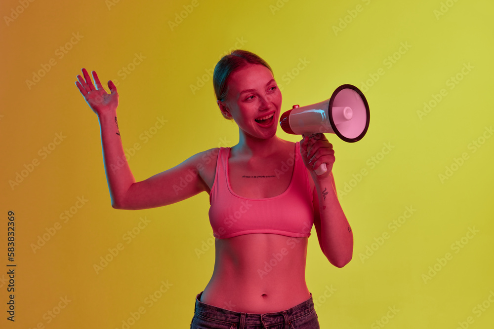 Emotional nice-looking girl telling something interesting through loudspeaker over yellow-orange background in neon light
