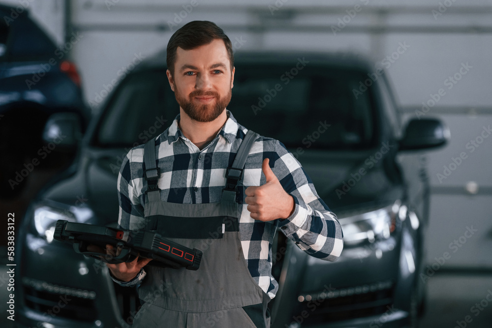 Showing gesture and smiling. Holding tablet. Man in uniform is working in the auto salon Showing gesture and smiling. Holding tablet. Man in uniform is working in the auto salon