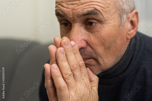 Please, Lord. Nervous worried aged Caucasian man praying on couch at home. Stressed white European senior citizen sitting on sofa, begging for forgiveness or asking God for help in difficult situation