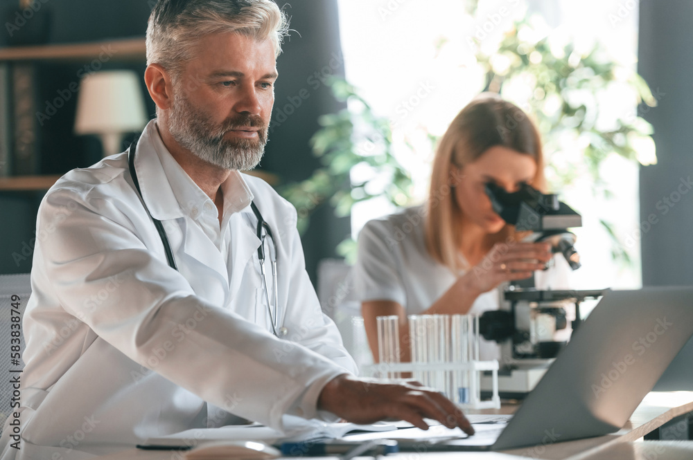 Using microscope. Two clinic workers are indoors together. Senior man with young woman Using microscope. Two clinic workers are indoors together. Senior man with young woman
