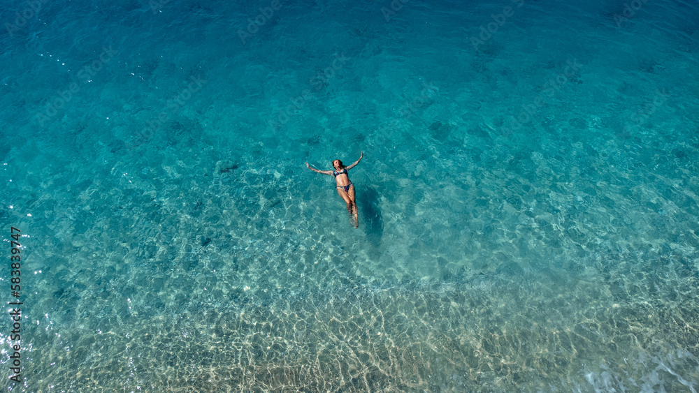 Aerial view of beautiful happy woman in swimsuit laying in the shallow sea water enjoying beach and soft turquoise ocean wave. Tropical sea in summer season on Egremni beach on Lefkada island.