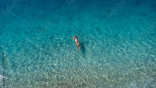 Aerial view of beautiful happy woman in swimsuit laying in the shallow sea water enjoying beach and soft turquoise ocean wave. Tropical sea in summer season on Egremni beach on Lefkada island. © MexChriss