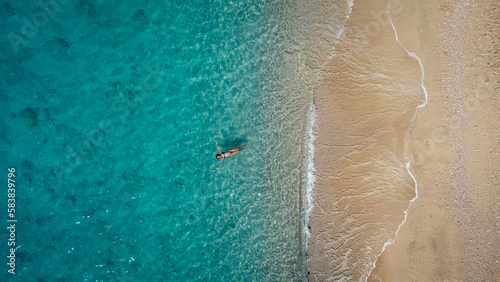 Aerial view of beautiful happy woman in swimsuit laying in the shallow sea water, enjoying sandy beach and soft turquoise ocean wave. Tropical sea in summer season on Egremni beach on Lefkada island.