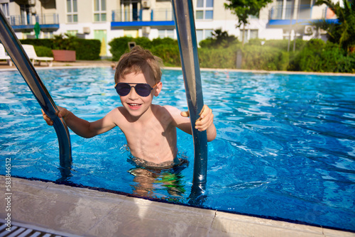 Obraz na plátně After swimming in the outdoor pool, the boy climbs out of the water up the steps, holding on to metal handrails