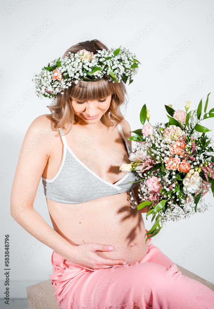 Pregnant woman in wreath with bouquet of flowers. Girl in gray top, pink pants. Hands on naked belly. Photoshoot happy pregnancy,maternity,preparation. Baby expectation. Beautiful tender young mother Pregnant woman in wreath with bouquet of flowers. Girl in gray top, pink pants. Hands on naked belly. Photoshoot happy pregnancy,maternity,preparation. Baby expectation. Beautiful tender young mother