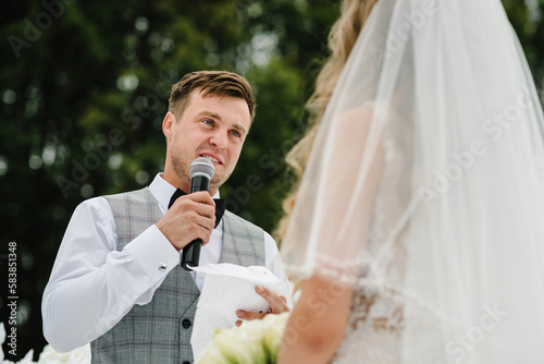Happy bride and groom exchanging wedding vows on ceremony under the arch on the backyard banquet area. The groom says the promise in the microphone. Fidelity in love.