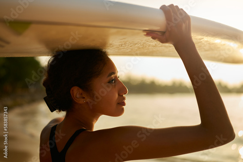 African american woman standing with surfboard on her head on ocean beach. Black female surfer posing with surf board. Pretty multiethnic girl goes on surfing session at sunny sunrise, enjoys sun.