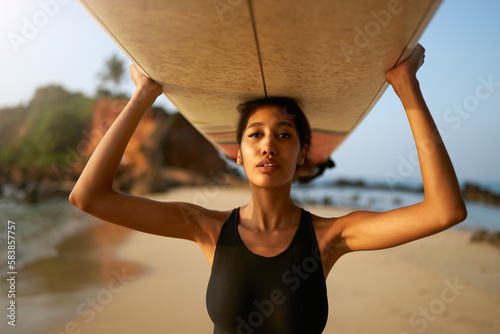 African american woman standing with surfboard on head on ocean beach. Black female surfer posing with surf board. Pretty multiethnic girl goes on surfing session on tropical location.