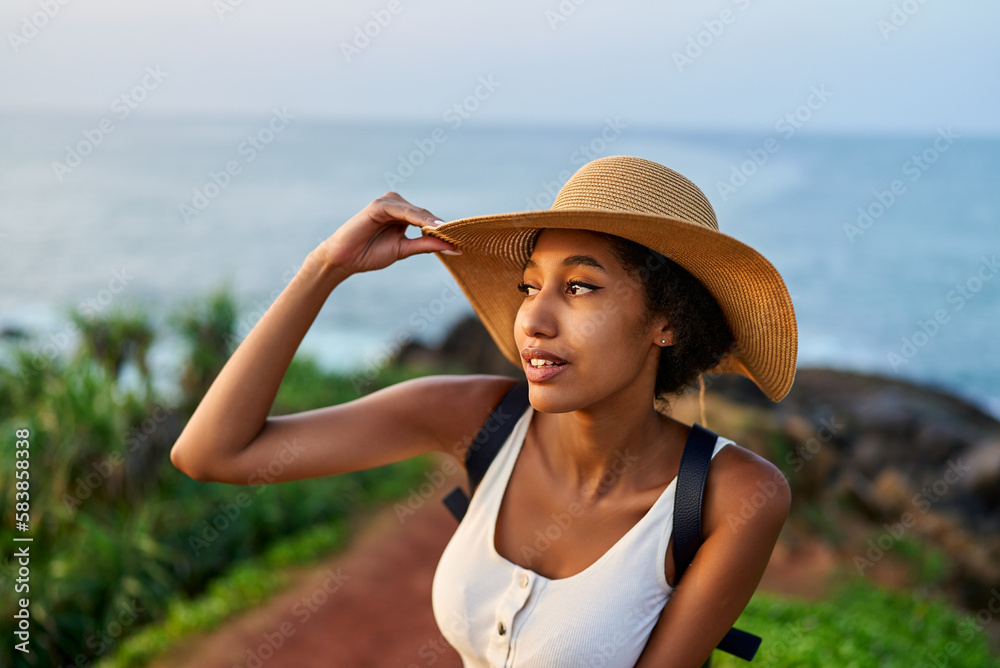 Multiethnic woman in straw hat enjoys tropical vacation travel hike. Black female with backpack sightseeing on scenic location. Pretty lady standing on island on sunrise. Girl in the sun for suntan