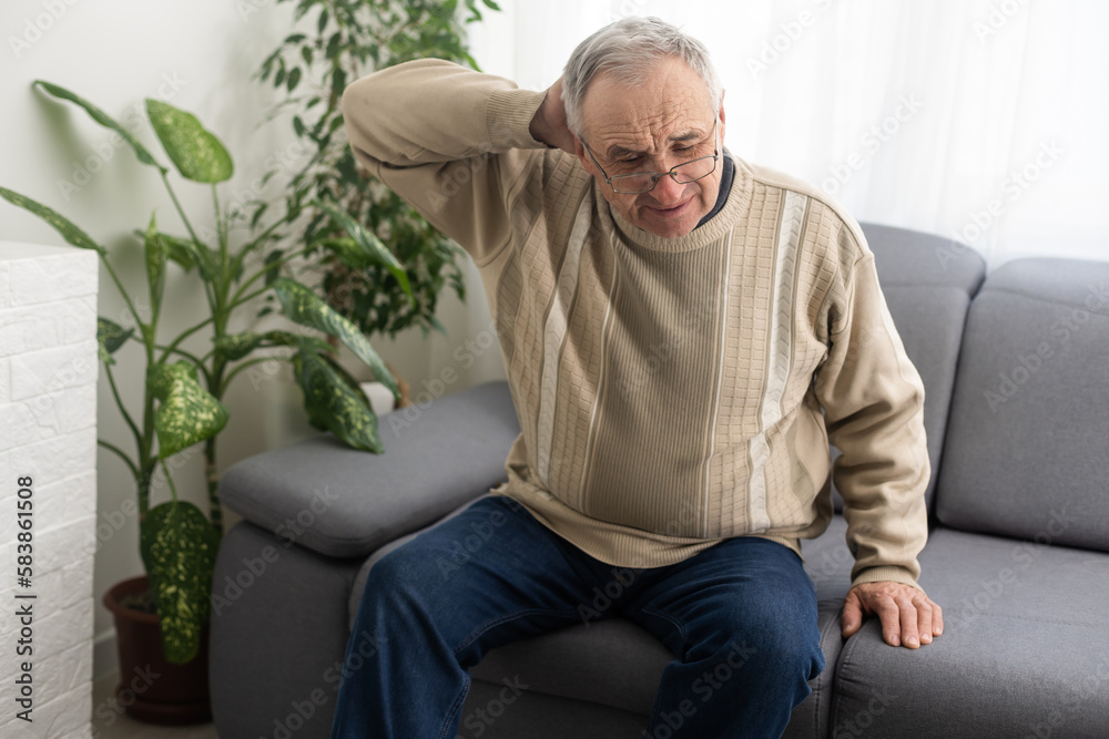Fototapeta premium Senior man having headache and touching his head while suffering from a migraine in the living room, Mature man presses a hand to head suffers from unbearable pain.