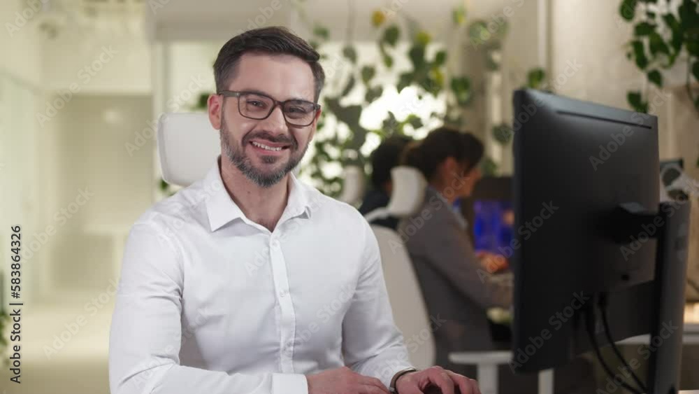 Portrait of happy bearded caucasian professional engineer working on computer typing on keyboard looking at camera smiling. Young cheerful successful man worker of IT company sitting in office. Portrait of happy bearded caucasian professional engineer working on computer typing on keyboard looking at camera smiling. Young cheerful successful man worker of IT company sitting in office.