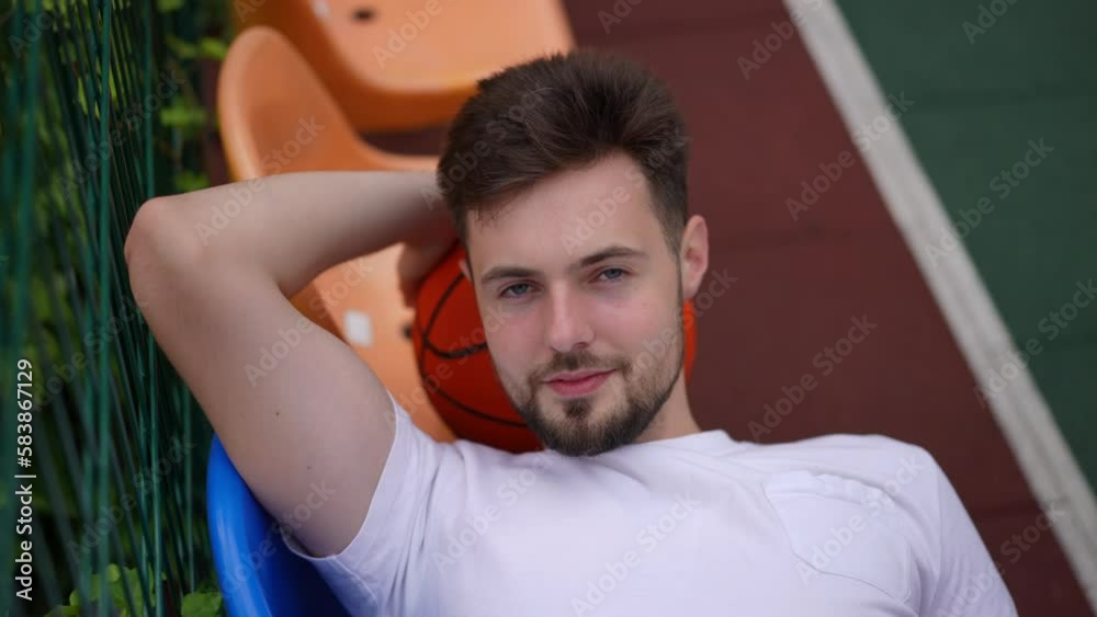 Top view young fit sportsman lying on chairs with basketball ball under head turning looking at camera in slow motion. Portrait of smiling satisfied confident motivated Caucasian man posing outdoors Top view young fit sportsman lying on chairs with basketball ball under head turning looking at camera in slow motion. Portrait of smiling satisfied confident motivated Caucasian man posing outdoors
