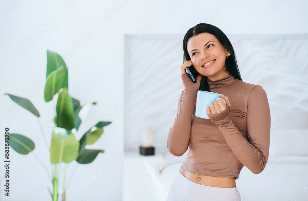 Happy Hispanic young brunette woman talks by phone holds cup of coffee at home looks happily away. Grateful European female businesswoman enjoying sunny morning. Successful people.