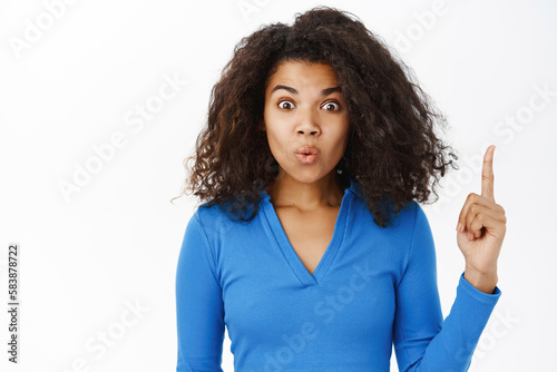 Portrait of curly woman pointing fingers up, showing advertisement on top, recommending click on link, demonstrating promo banner, wearing blue top