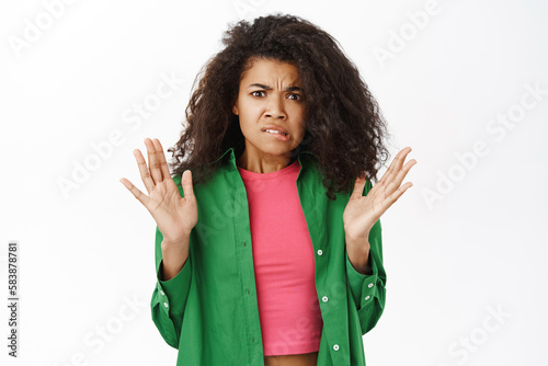 Portrait of disgusted woman raise hands up, wrinkles nose and looks freaked out, dislike smth nasty, standing over white background photo