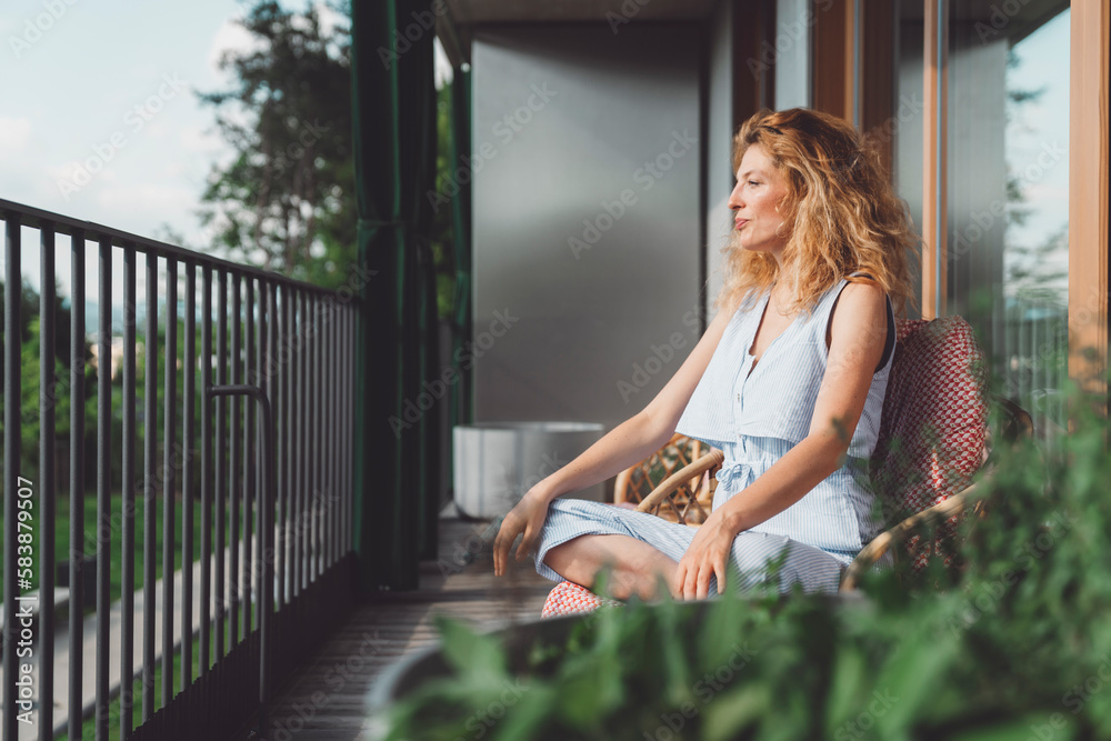 Side view of a woman relaxing on the balcony sitting in an armchair looking out in the nature Side view of a woman relaxing on the balcony sitting in an armchair looking out in the nature