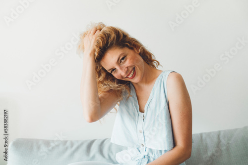 Waist up portait beautiful woman sitting on the couch playing with her hair smiling looking at the camera