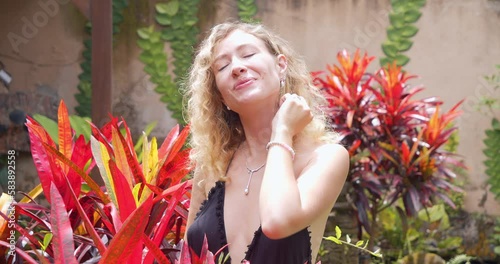 Close-up portrait of a young smiling woman looking at the camera and enjoying the sun among the flowers in the garden