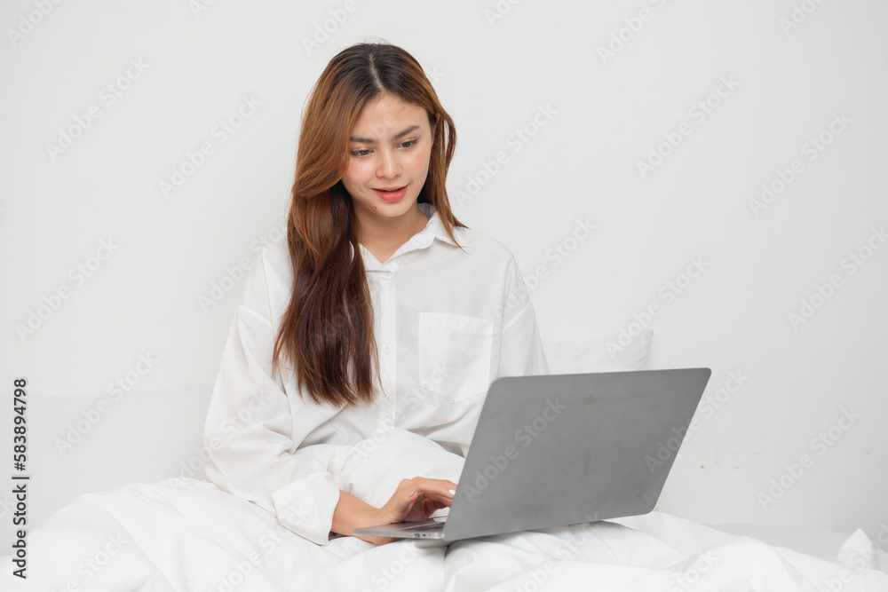 Woman in white nightgown waking up on weekend morning resting and relaxing playing with laptop mobile phone Eating bread and drinking tea in glass inside white bedroom. Morning vacation concept. Woman in white nightgown waking up on weekend morning resting and relaxing playing with laptop mobile phone Eating bread and drinking tea in glass inside white bedroom. Morning vacation concept.