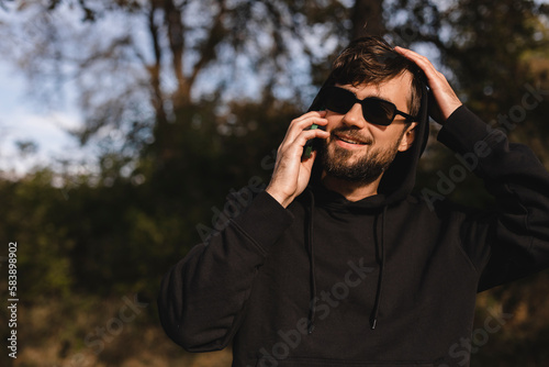 Handsome man have mobile phone conversation and fix his hair. Man wear black hoody and glasses.