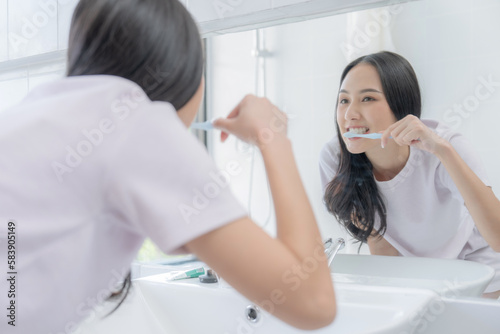 Smiling young Asian woman brushing teeth in bathroom.