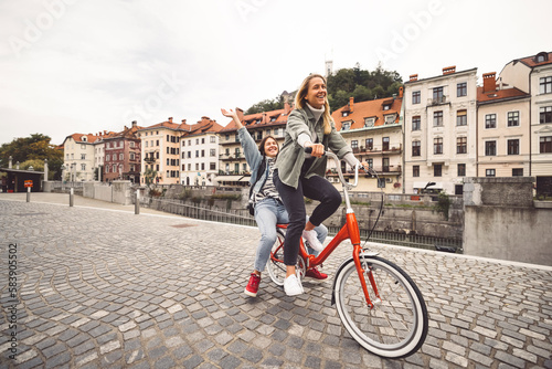 Two cheerful young women , best friends having fun in the city riding a red bike, one sitting on the drunk of the bike 