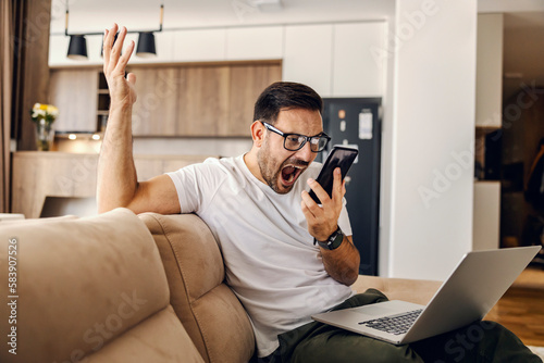 Foto An angry man sits at home on sofa and yelling at the phone while holding a laptop in his lap