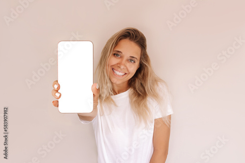 Young caucasian smiling blonde woman holds mobile phone in her hand demonstrating white screen with empty copy space for text or design on beige background. Smartphone mockup in the foreground closeup