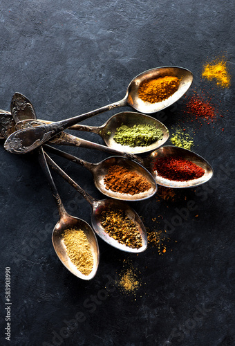Spices, herbs and seasonings in spoons on a black textured background, flat lay