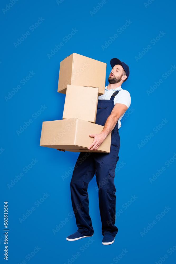 Full size body portrait of busy attractive deliver in blue uniform with stubble, having three heavy big boxes in arms, isolated on grey background, trying to withhold Full size body portrait of busy attractive deliver in blue uniform with stubble, having three heavy big boxes in arms, isolated on grey background, trying to withhold