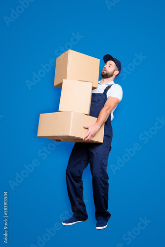 Full size body portrait of busy attractive deliver in blue uniform with stubble, having three heavy big boxes in arms, isolated on grey background, trying to withhold