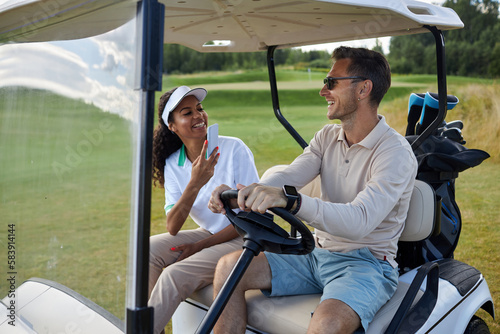 Portrait of multiethnic sporty couple taking photos in golf cart outdoors and smiling cheerfully