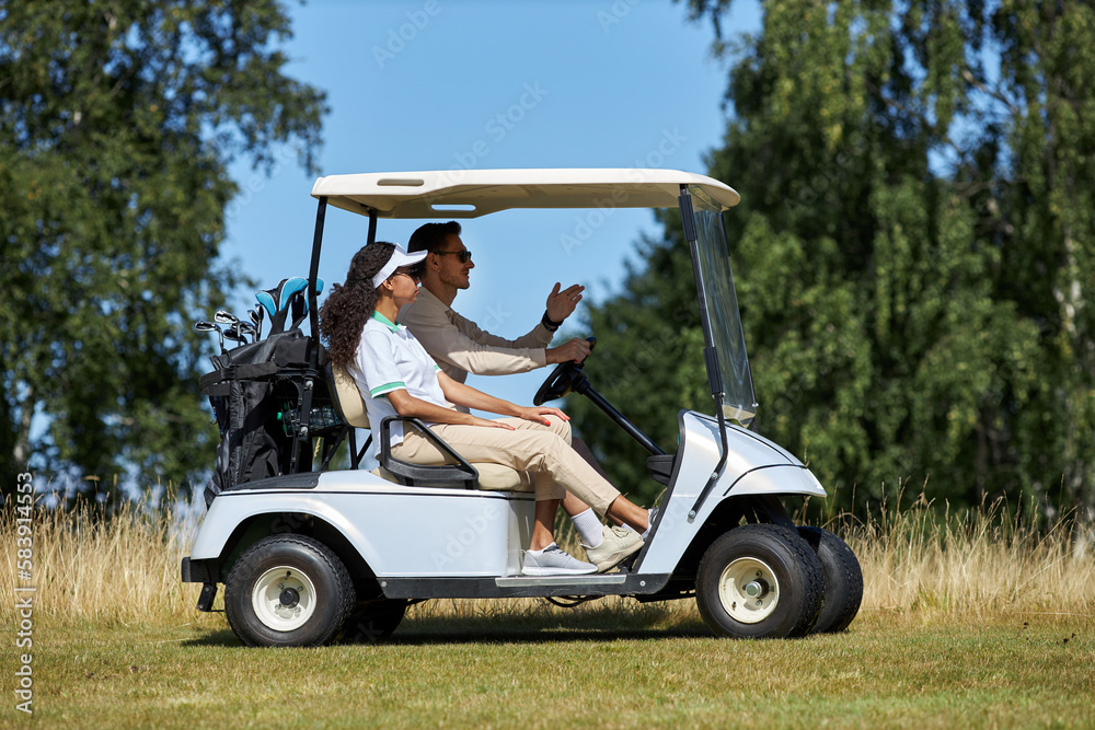 Side view portrait of sporty young couple driving golf cart across field in sports club lit by sunlight