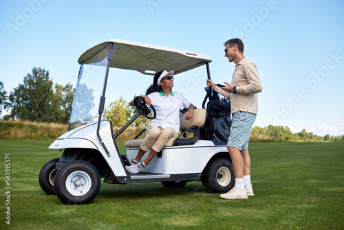Full length portrait of sporty young couple chatting by golf cart outdoors in green field