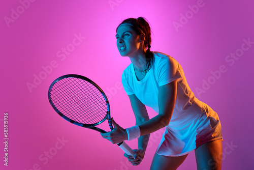 Concentrated young woman, professional female tennis player in white uniform posing with racket against pink studio background in neon light. Concept of professional sport, movement, health, action © master1305
