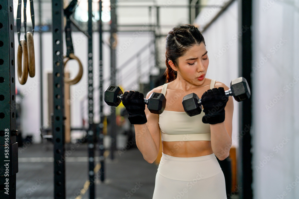 Pretty Asian sport woman hold and lift two dumbbells up and look to her hands in fitness gym and look concentrate to training.
