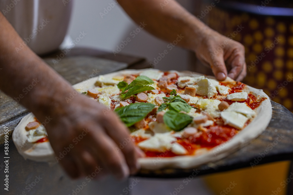 Pizza tradizionale napoletana condita con sugo di pomodoro, würstel, mozzarella di bufala e basilico fresco mentre viene stesa sulla pala di una pizzeria napoletana e pronta per essere infornata